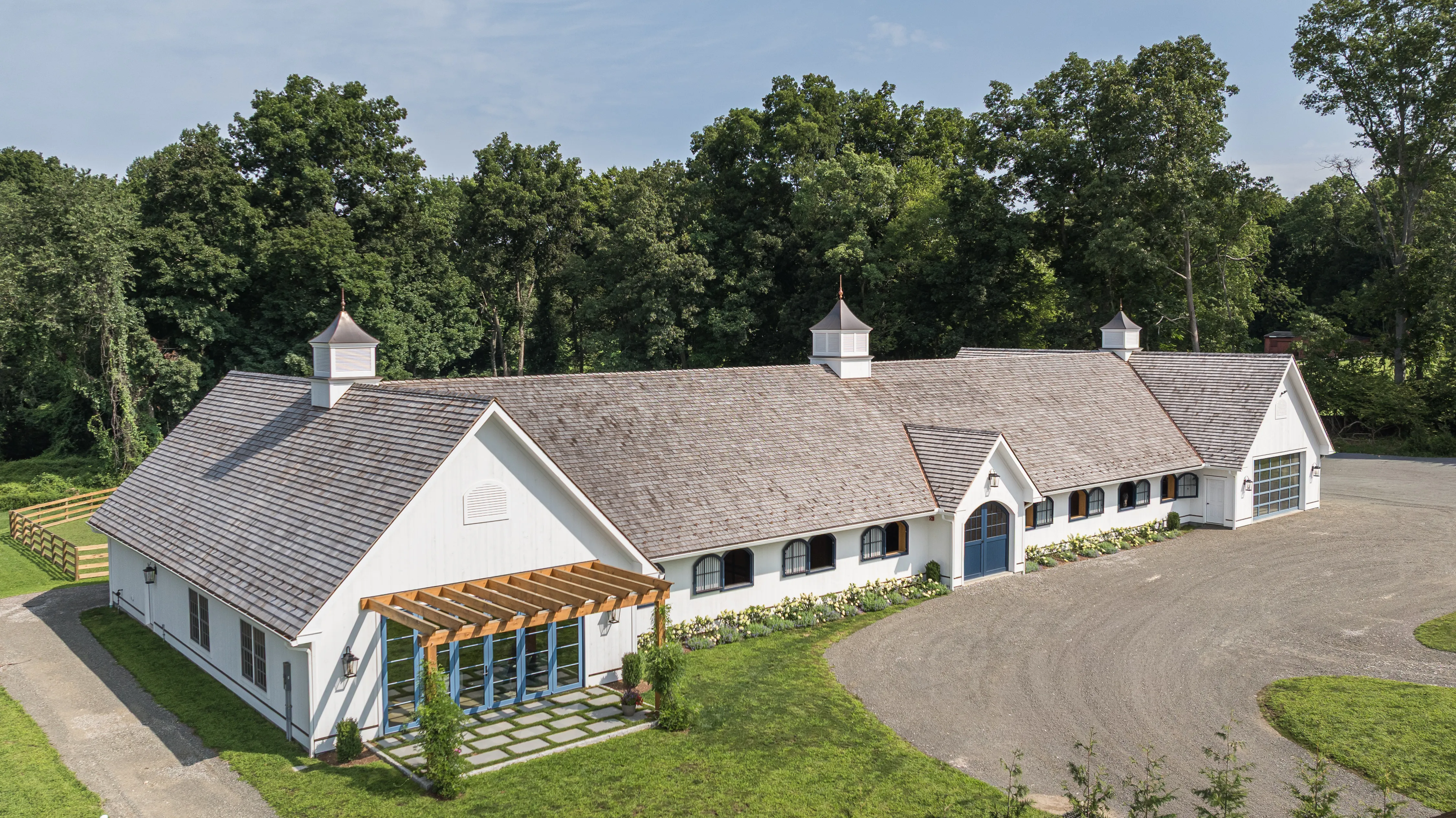 A white horse stable with three cupolas, a grand double-door entrance, and matching bay garage doors sits along the woods.