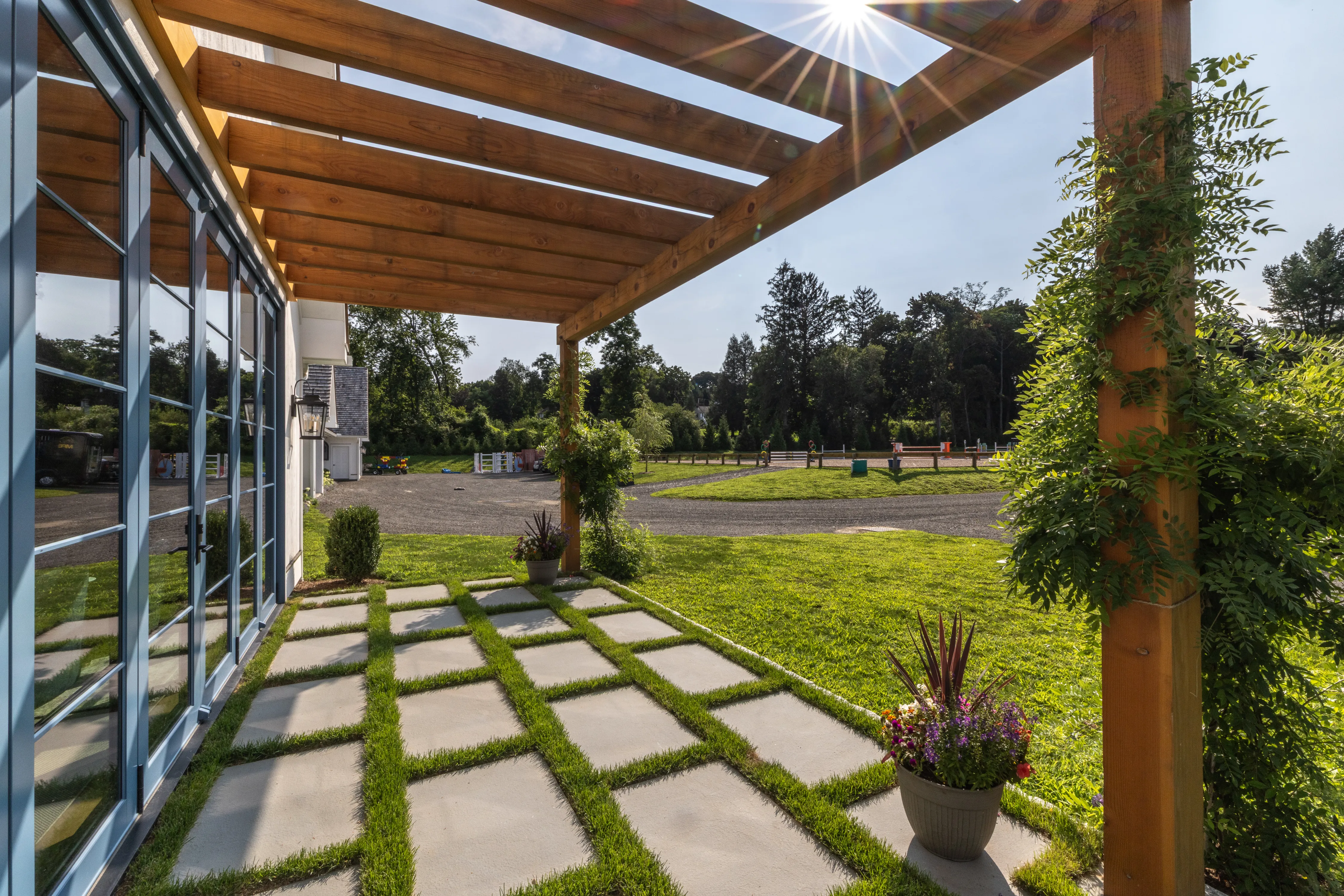 A timber frame pergola brings shade to an exterior stone patio that is framed by a neatly manicured yard and colorful plants.
