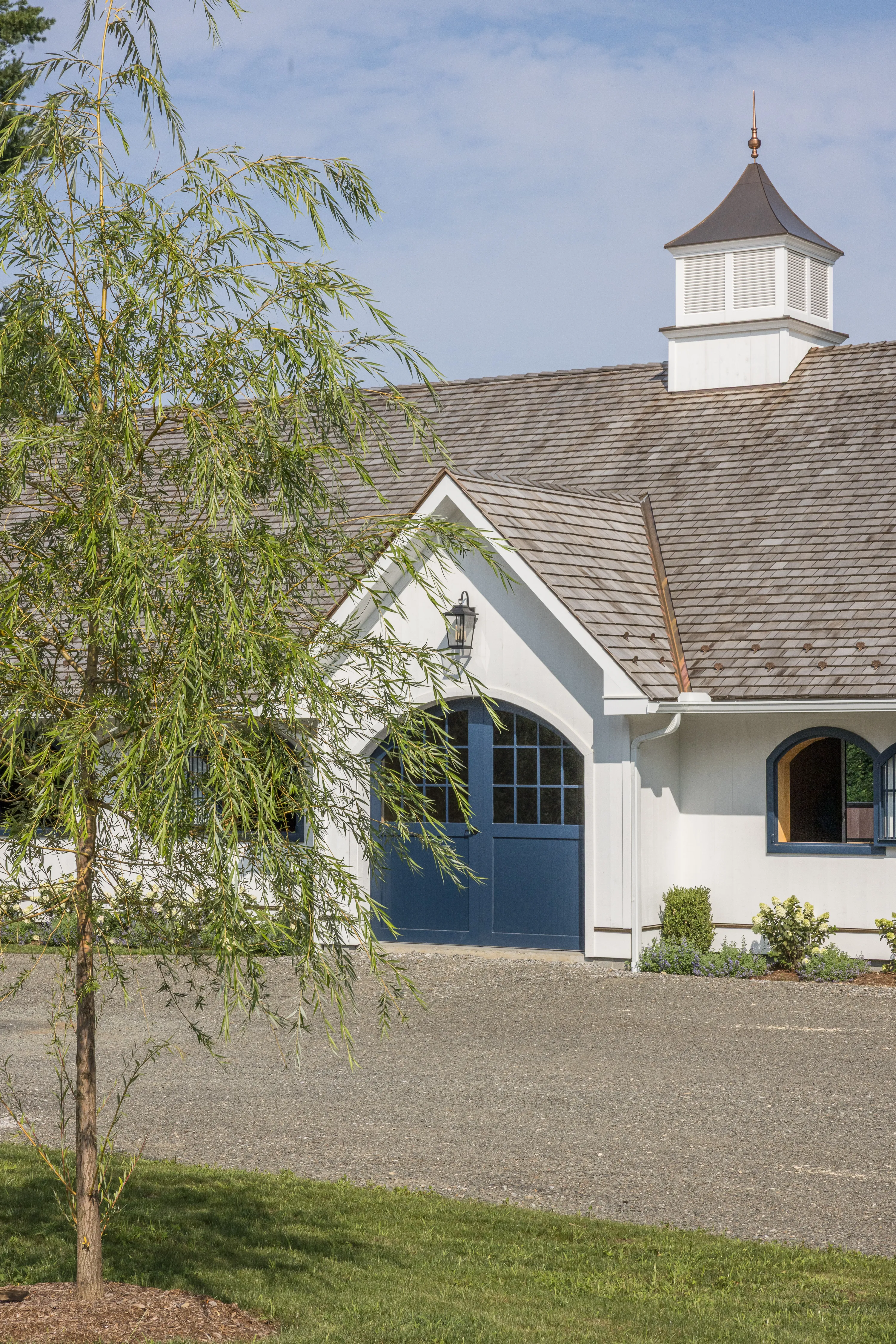 A tree sits in front of a white horse barn with blue doors and windows and a cedar shake roof in the background.