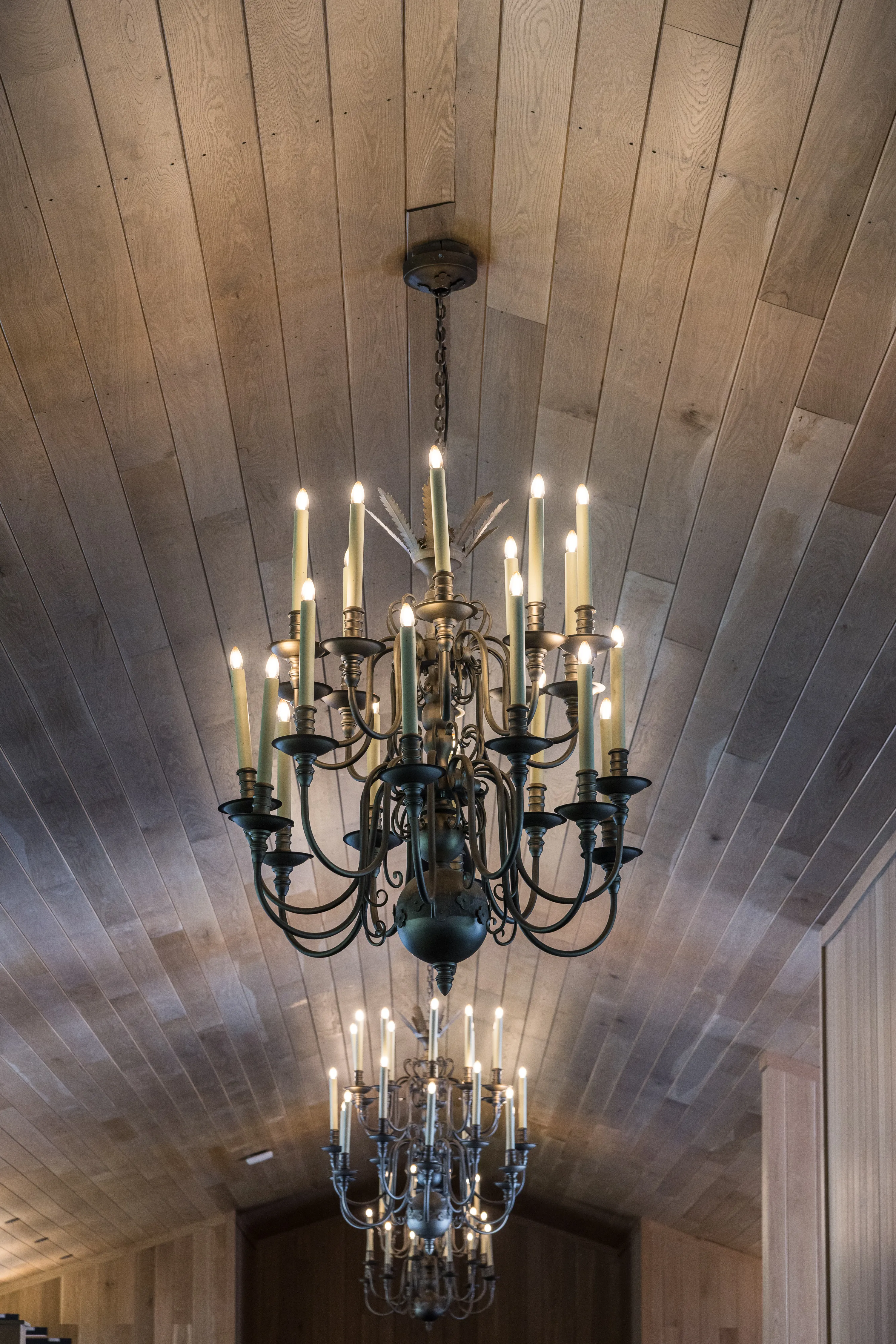 An ornate wrought-iron chandelier with candle-style lights hangs from a white oak-paneled horse barn ceiling.