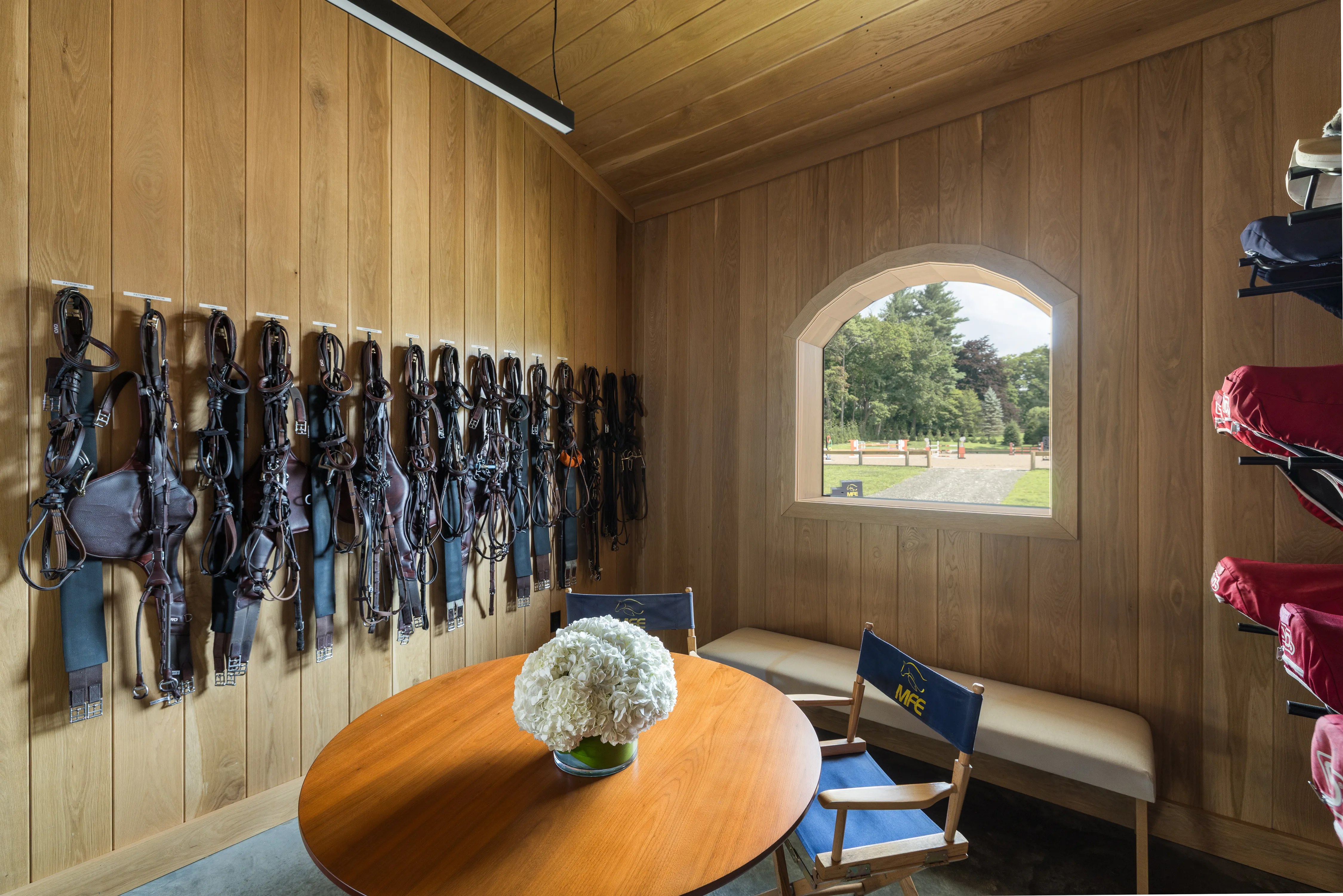 An orderly white oak tack room has tack hanging on the walls and abundant natural light coming through an arched window.