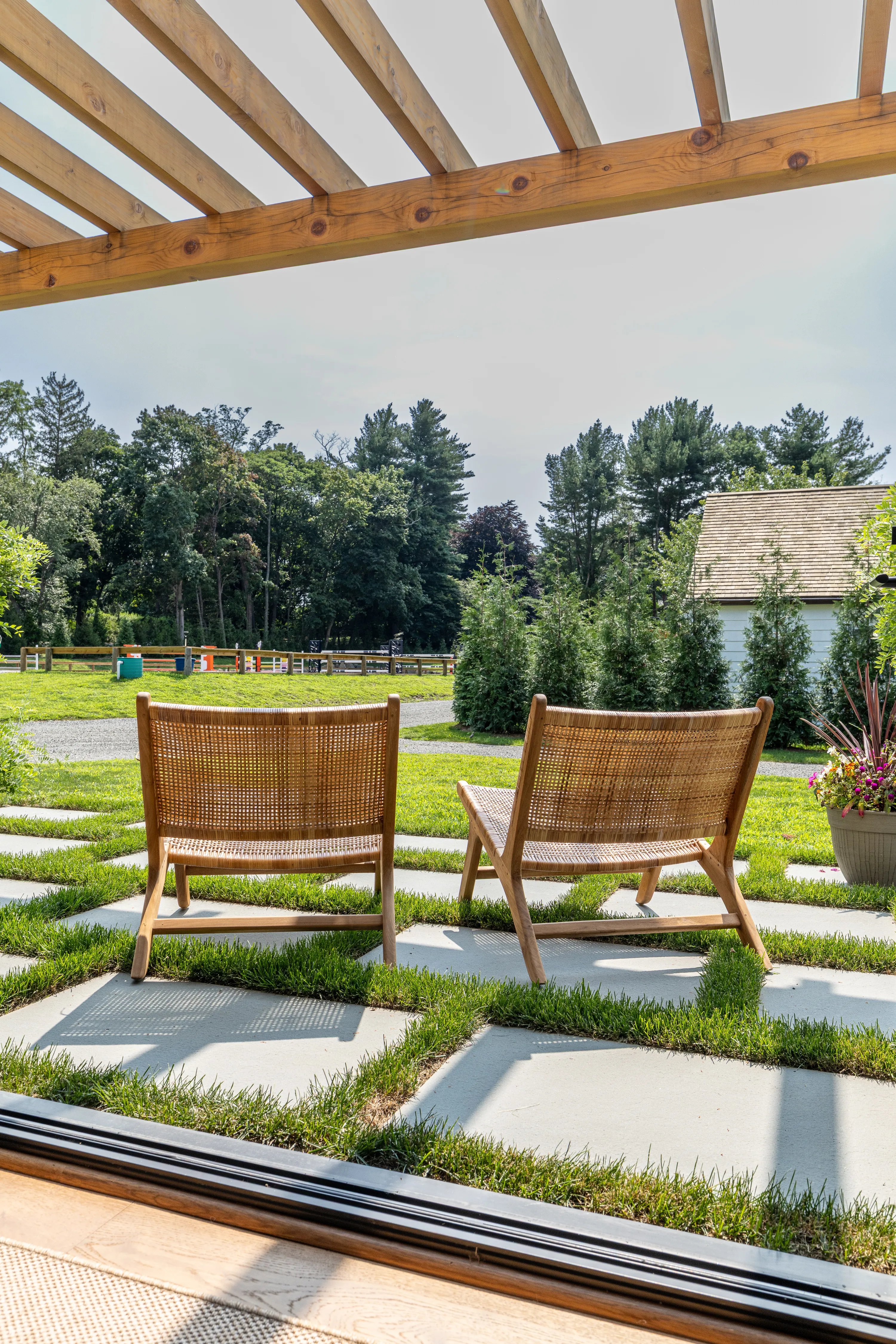 Two wooden chairs sit on the stone patio covered by a timber frame pergola, looking out over the horse barn’s estate.