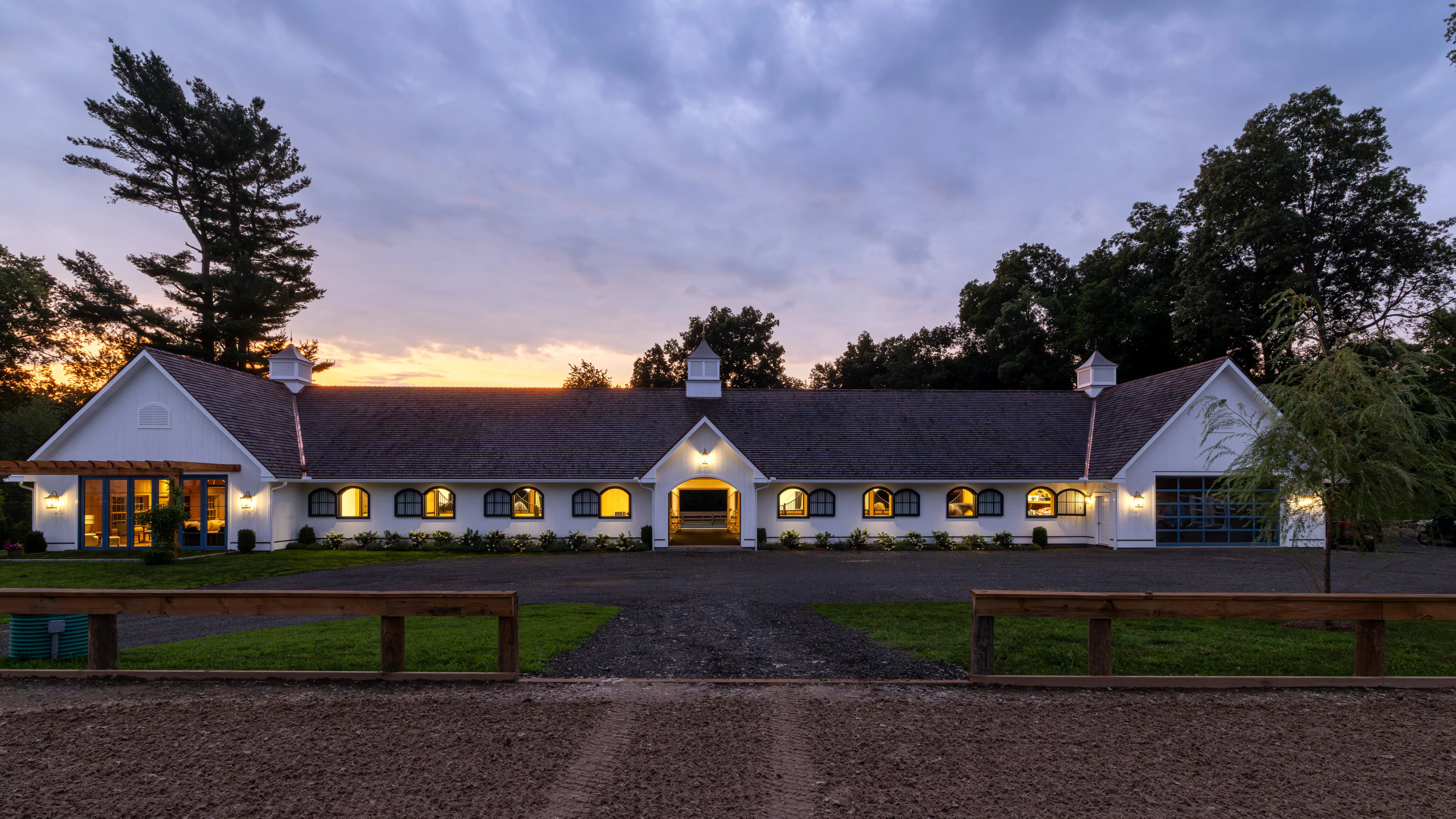 A twilight image of a white barn with interior light shining through the open windows while three cupolas adorn the roof.