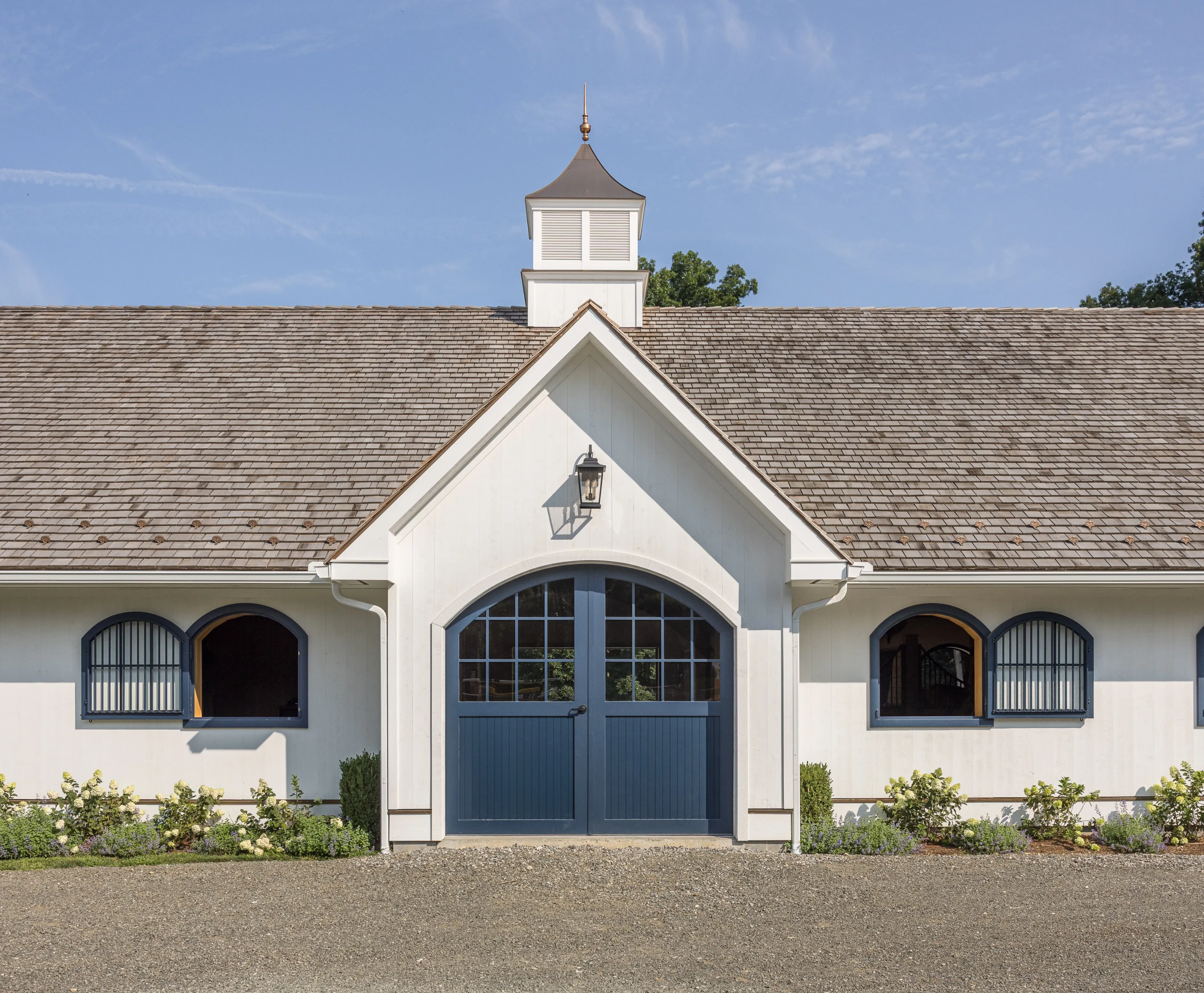 A blue double-door entrance leads into a horse barn with arched stall windows on either side and a black lantern sconce above.