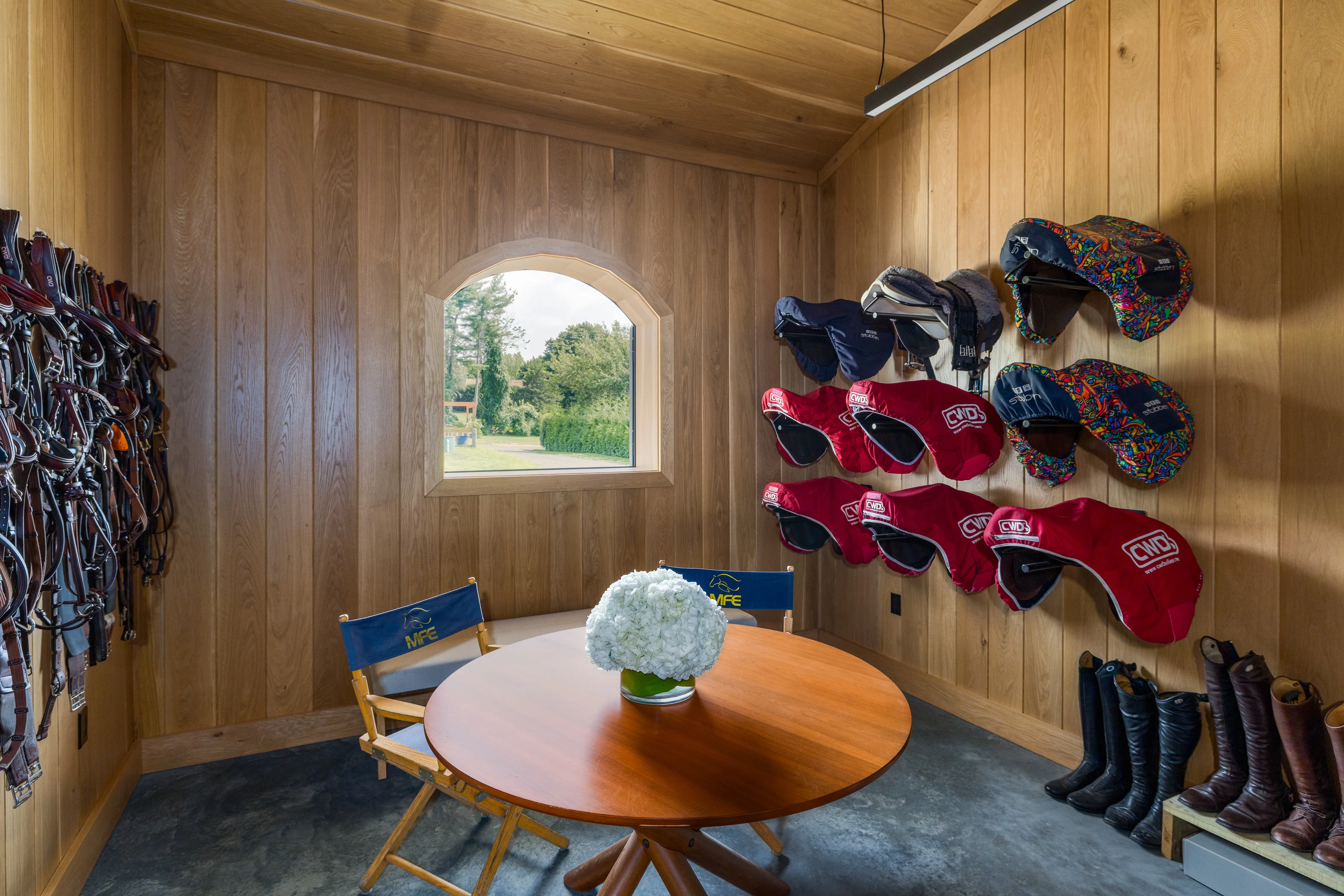 An orderly white oak tack room has saddles hanging on the walls and abundant natural light coming through an arched window.