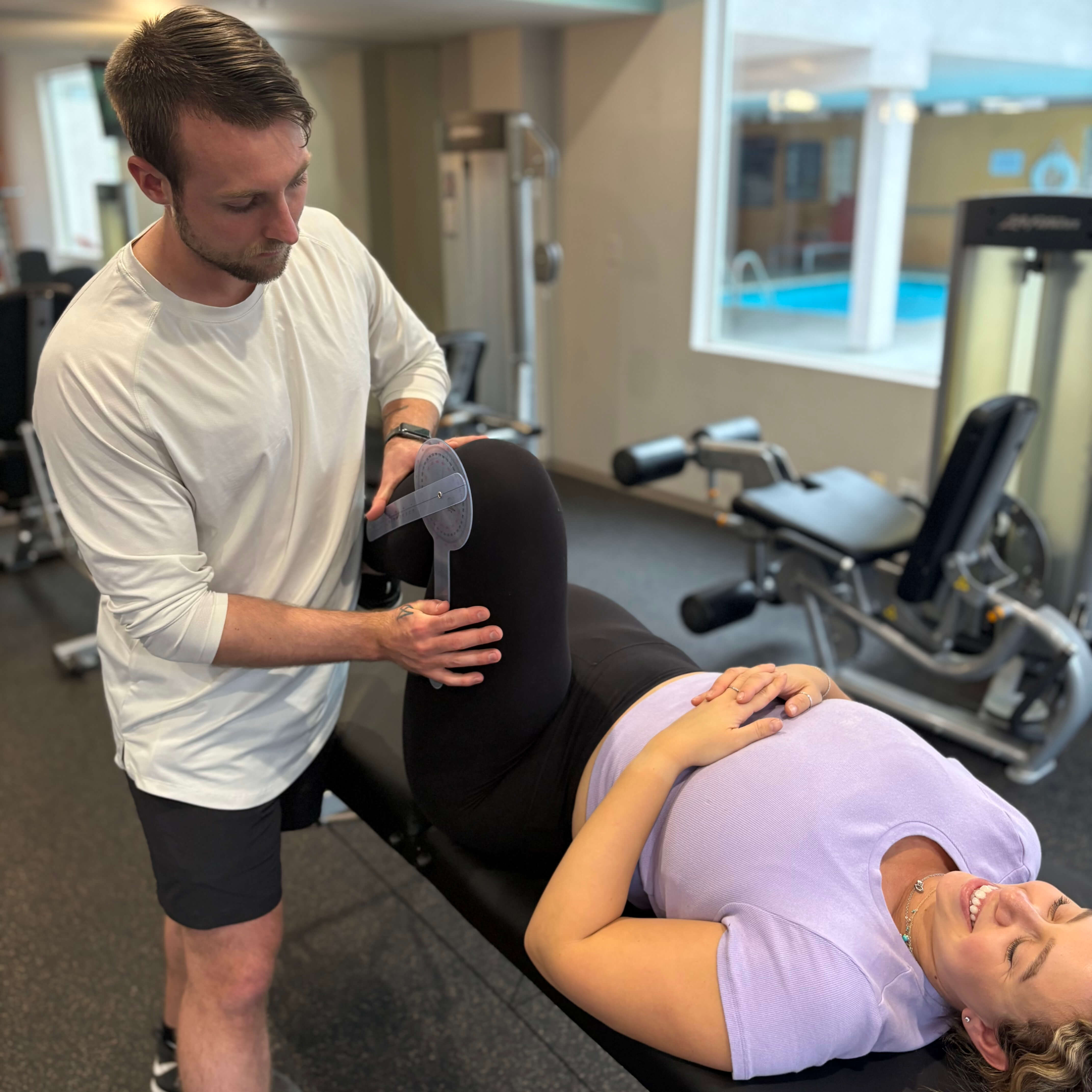 Physical therapist measuring knee angle of a smiling woman lying on a bench in a gym.