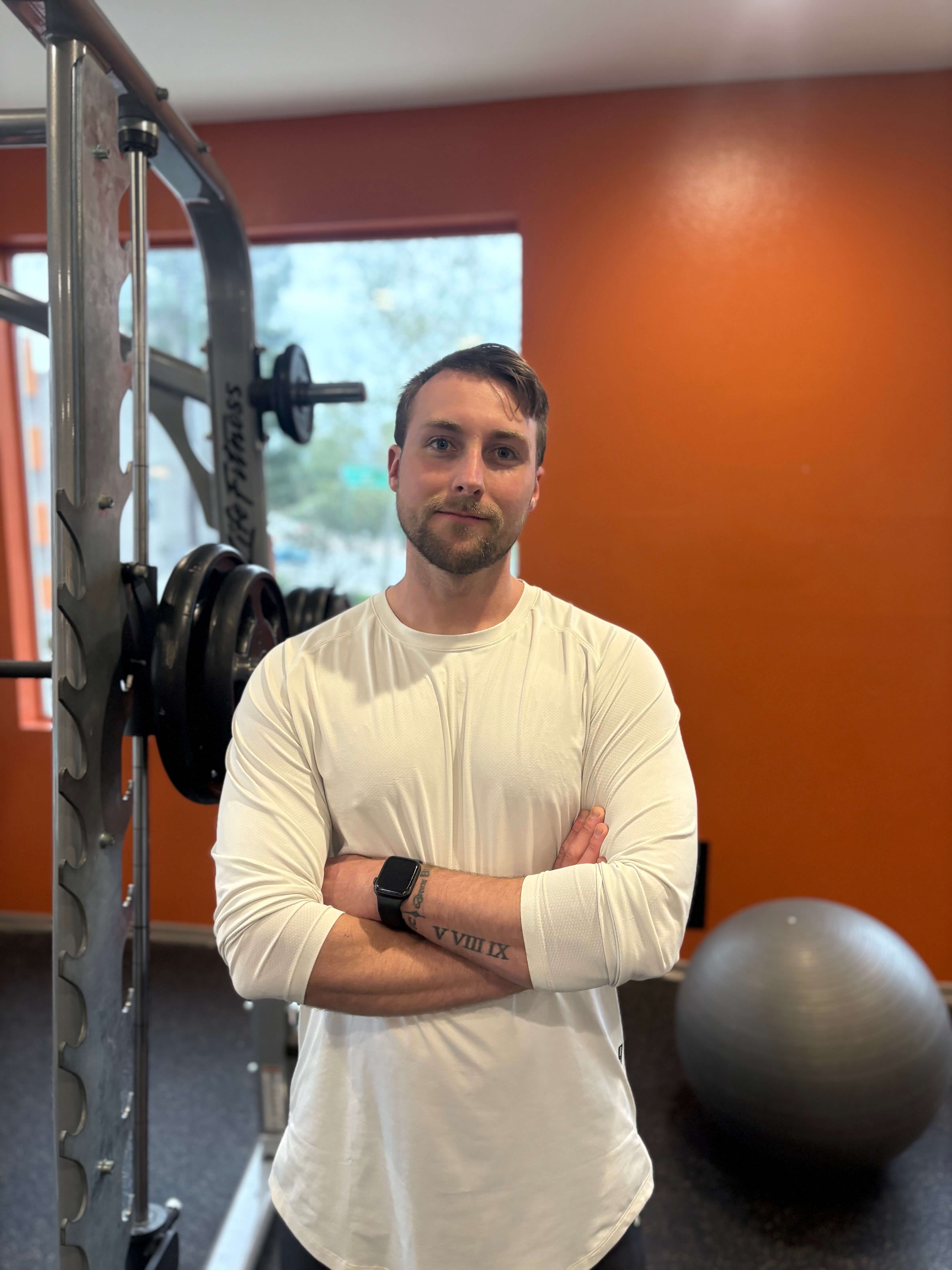 Man with folded arms wearing a white long-sleeve shirt and smartwatch standing in a gym with weightlifting equipment and an exercise ball.