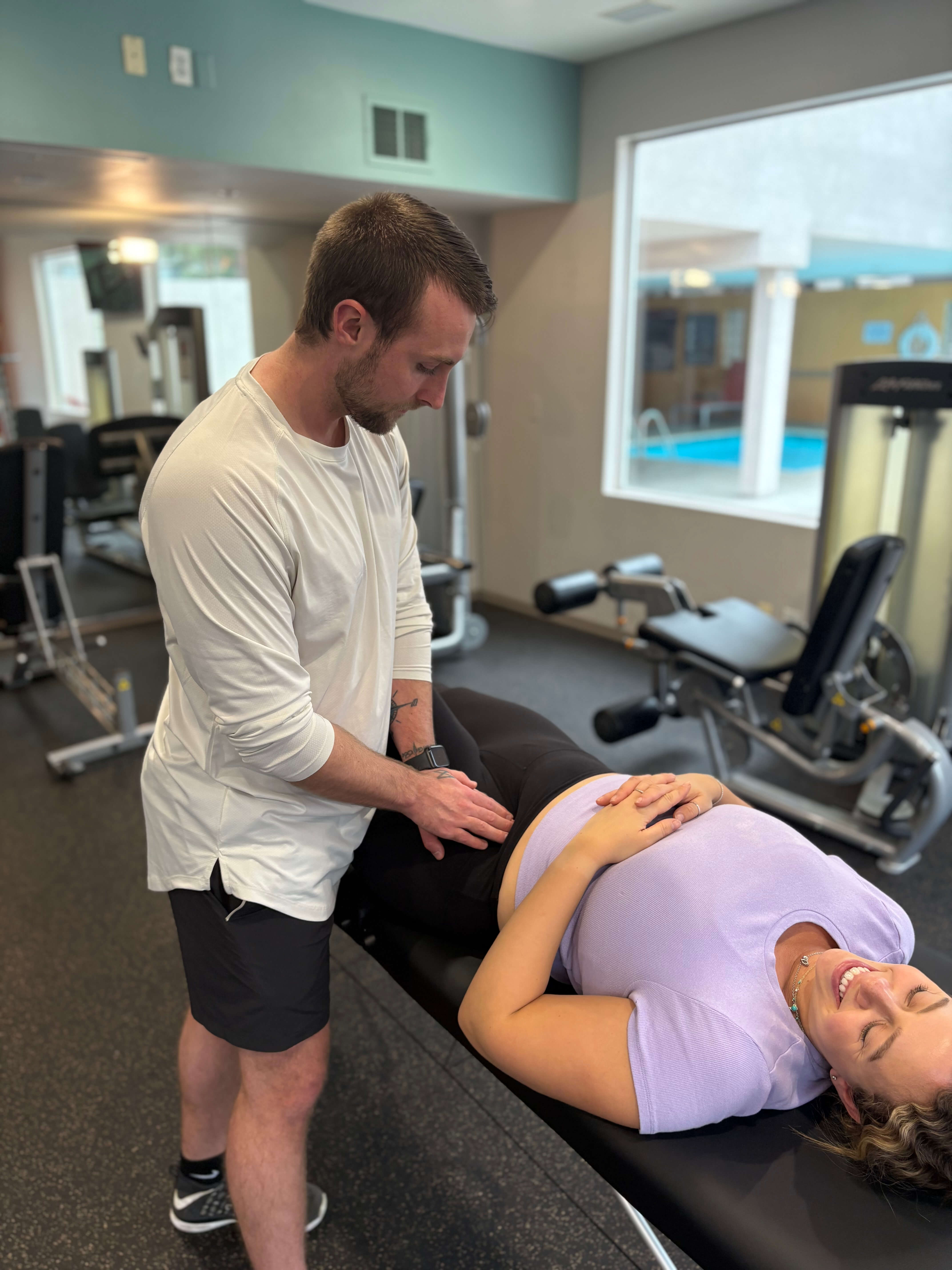 A man in a white shirt and black shorts is performing a physical therapy or chiropractic treatment on a smiling woman lying on a black treatment table in a gym setting.