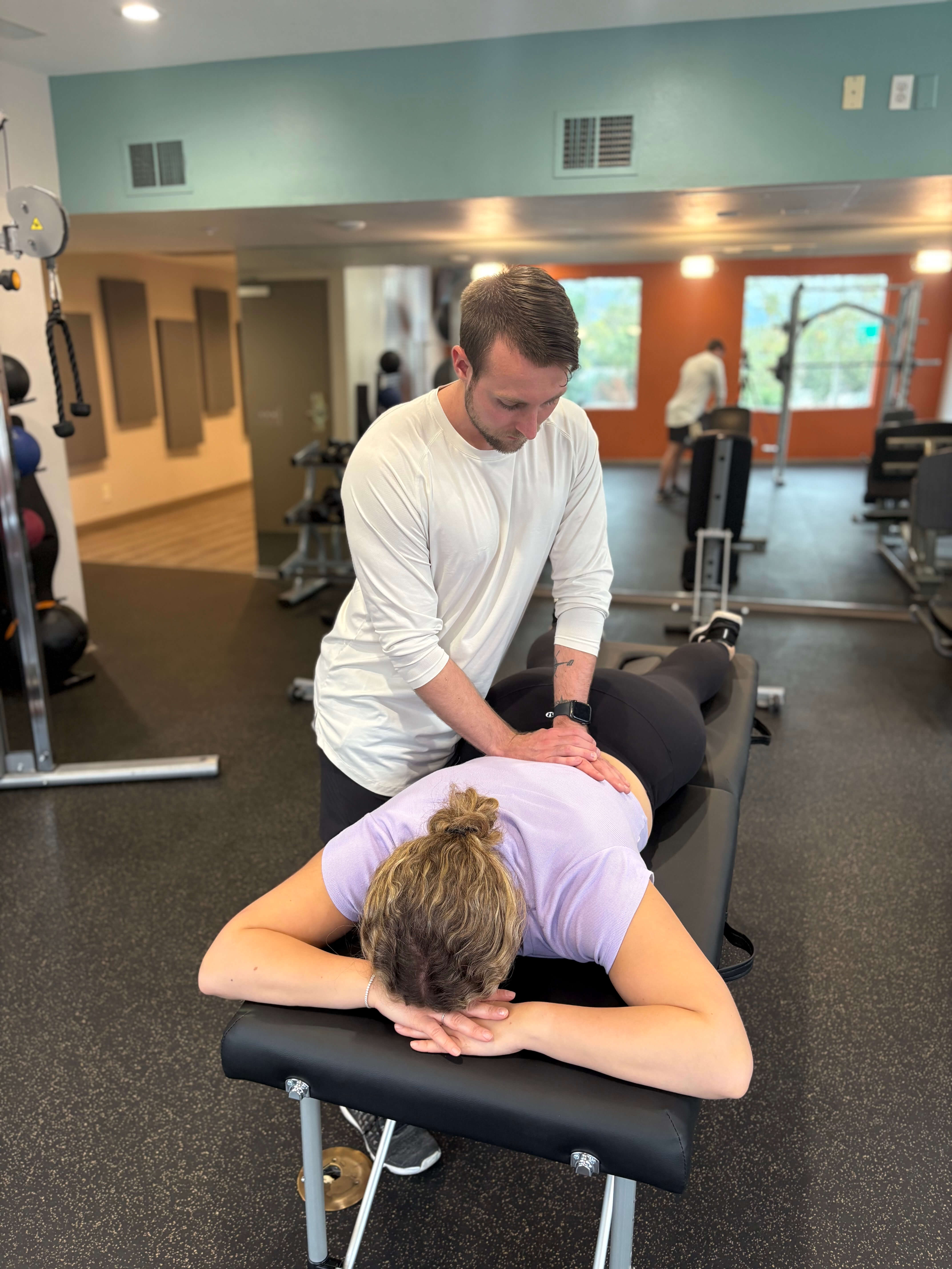 Therapist performing a back massage on a woman lying face down on a massage table in a gym.
