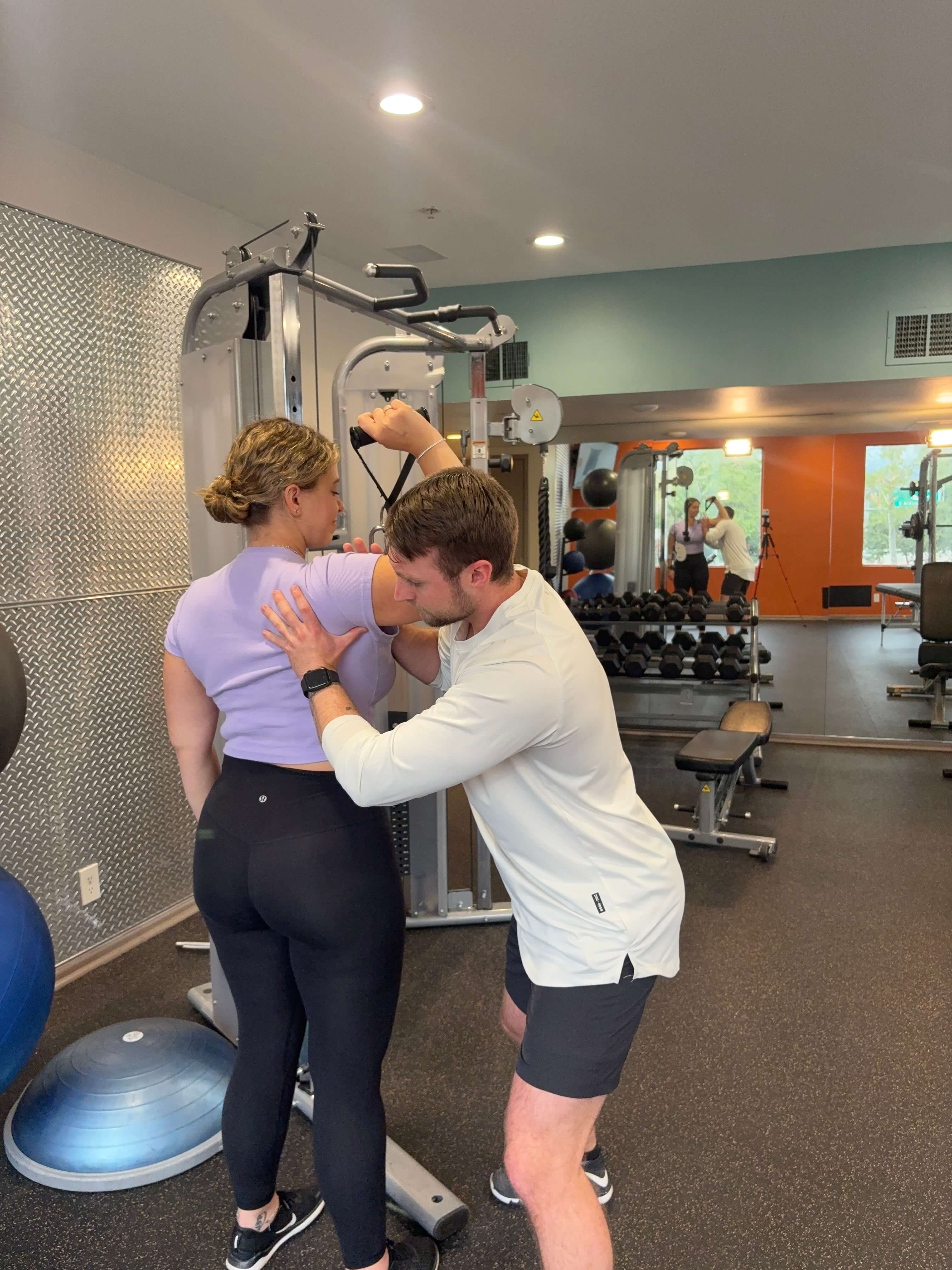 A male personal trainer assists a female client with a stretching exercise on a gym cable machine.