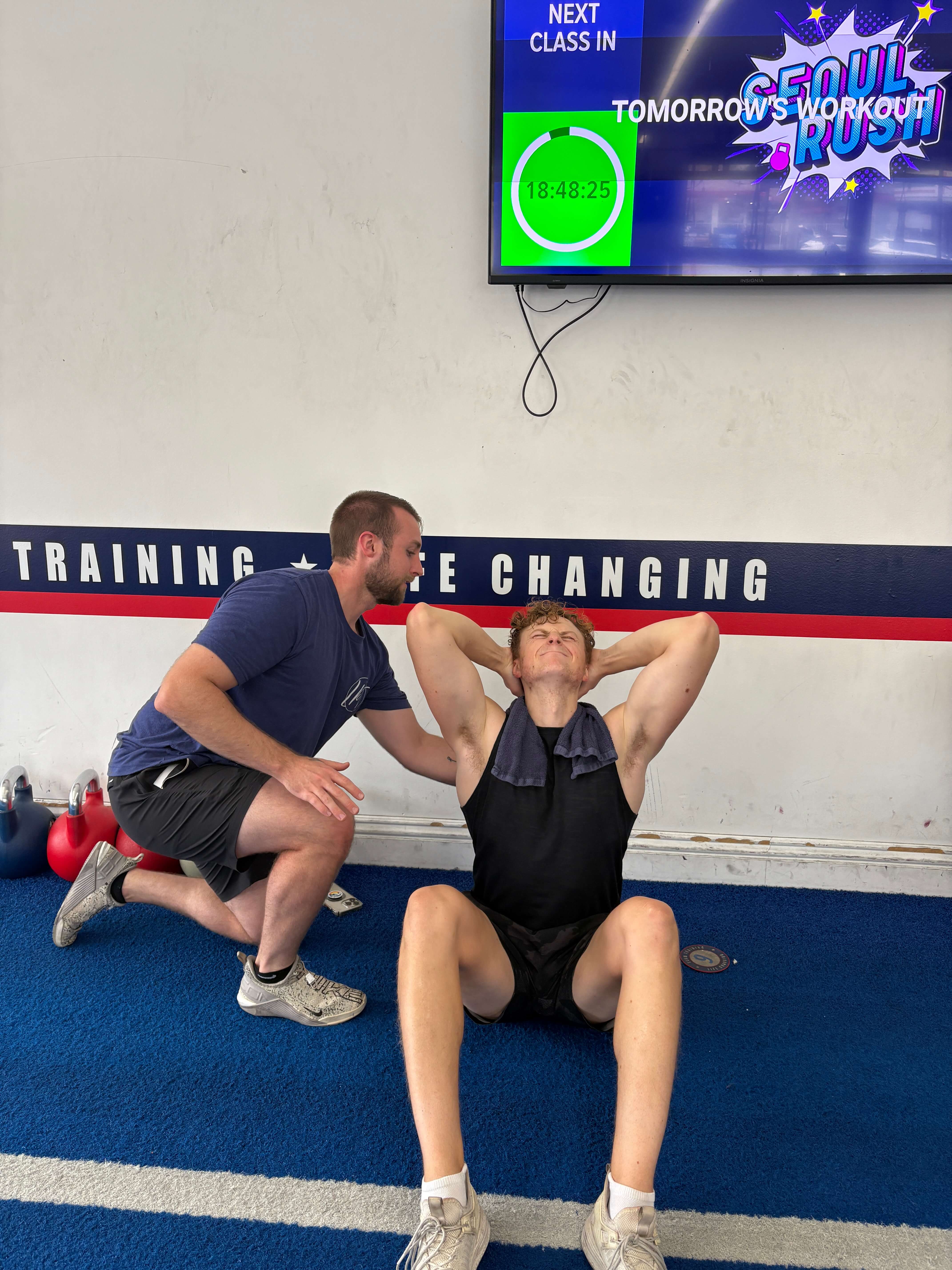 Trainer assists a man doing sit-ups in a gym with kettlebells and a timer on the wall showing 18:48:25.