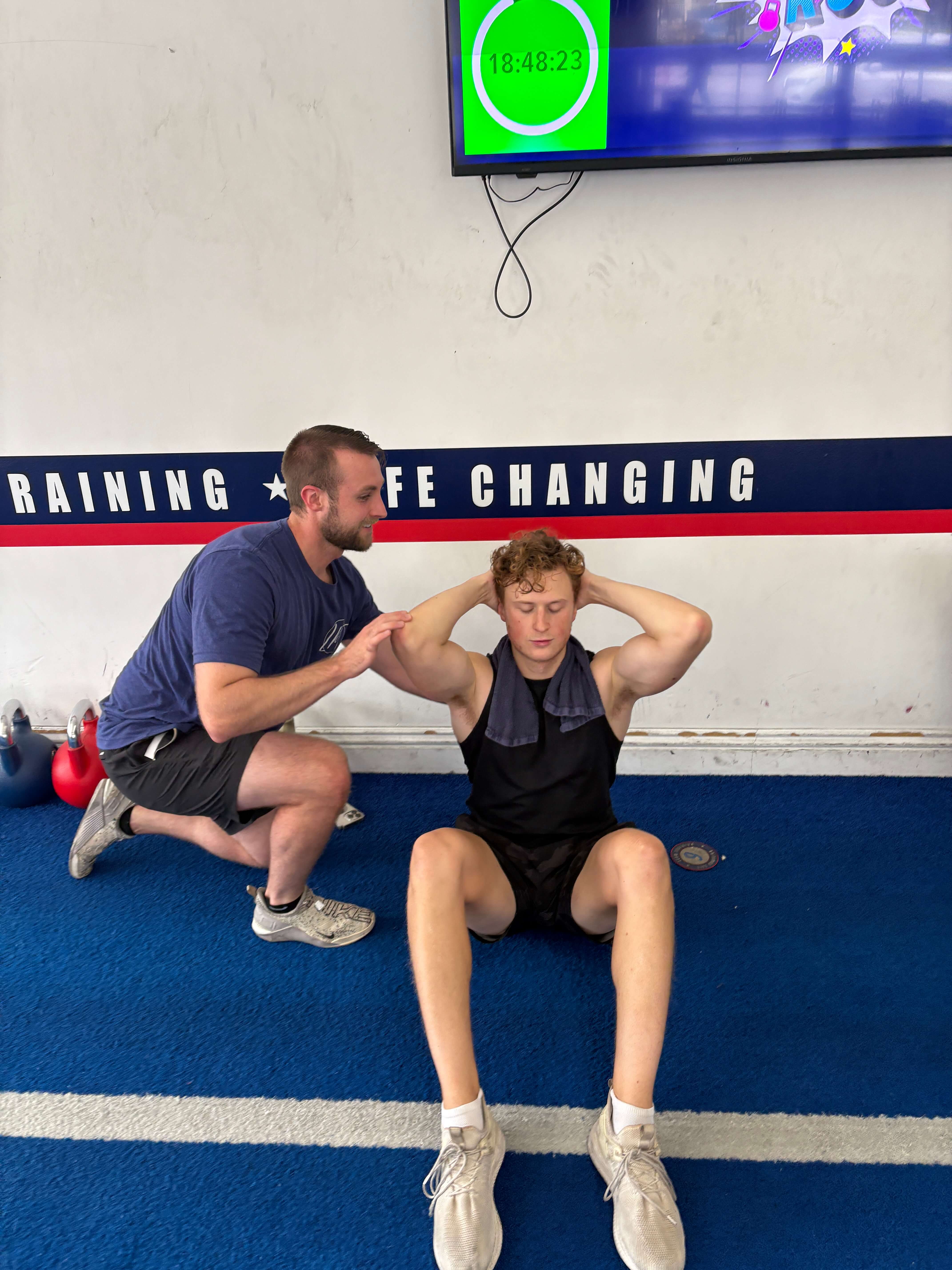 A personal trainer kneeling and coaching a young man doing sit-ups on a blue gym floor.