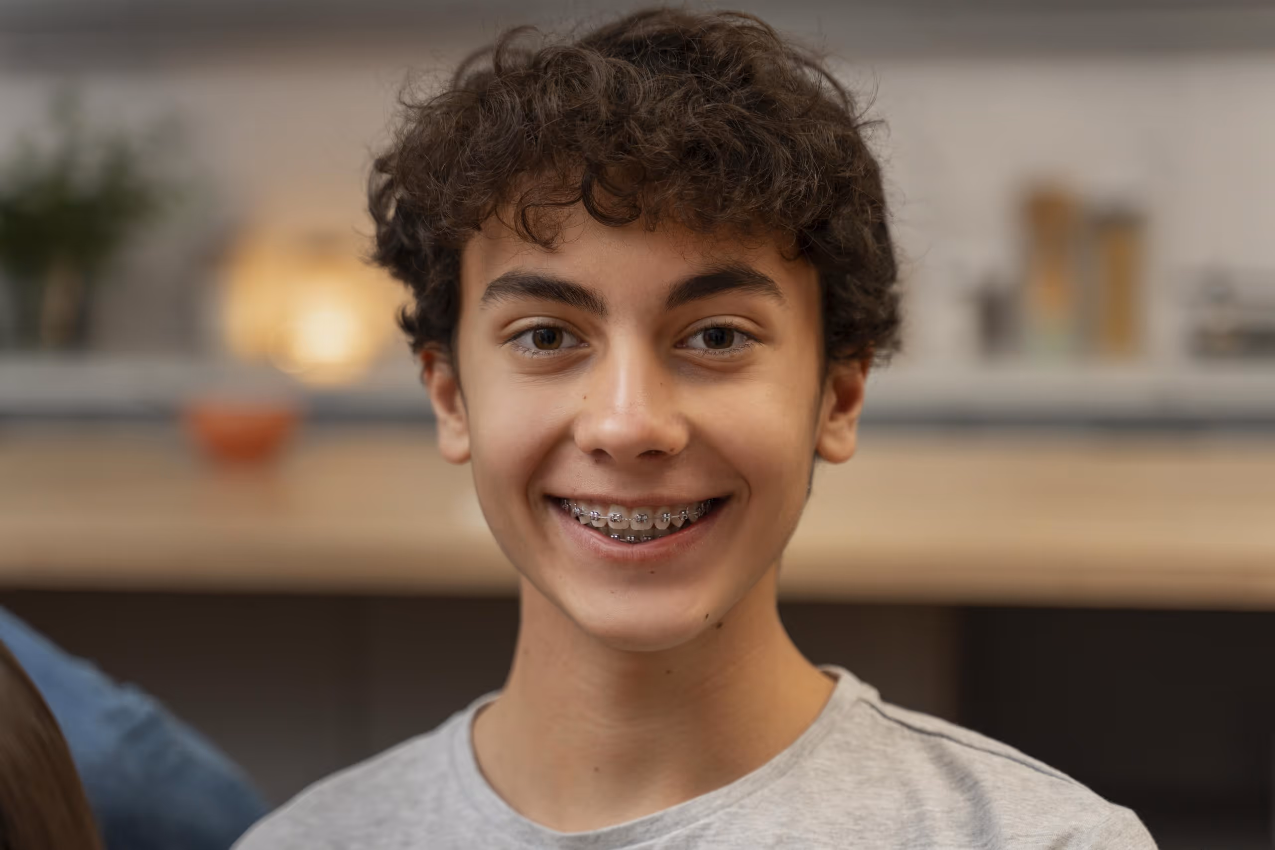 Smiling teenage boy with curly hair and braces wearing a light gray shirt against a blurred indoor background.
