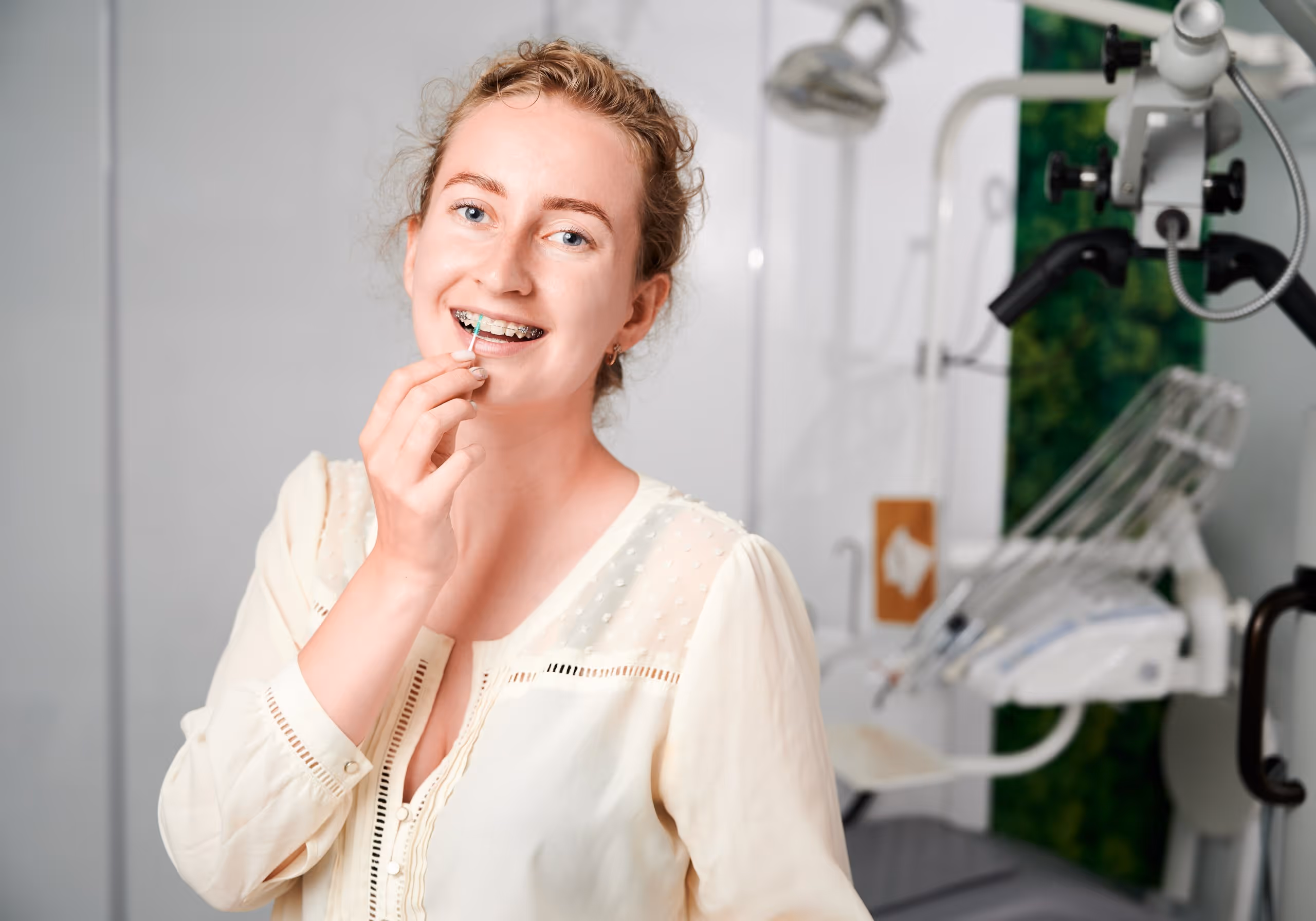 Smiling woman with braces cleaning her teeth with an interdental brush in a dental office.