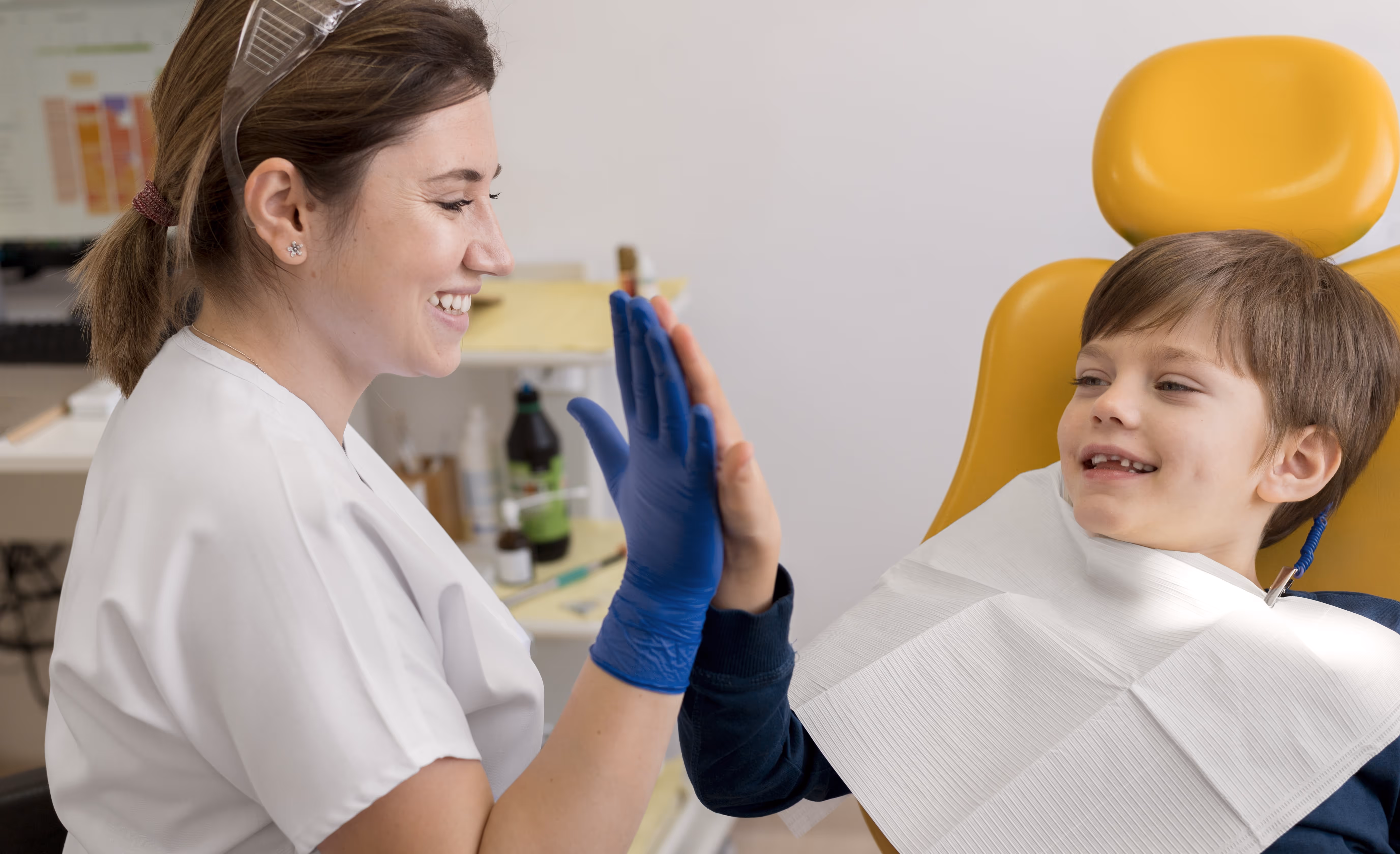 Smiling female dentist in white coat gives a high-five to a happy young boy sitting in a yellow dental chair with a bib.