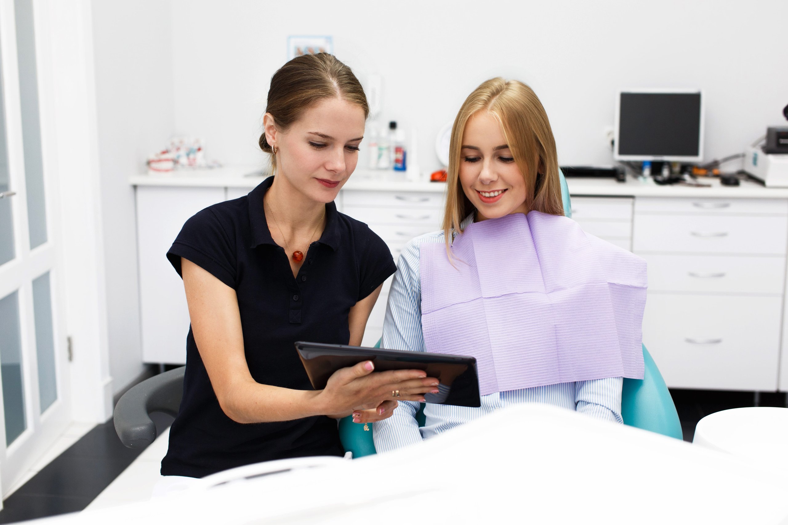 Dentist showing a tablet to a female patient sitting in a dental chair with a purple bib.