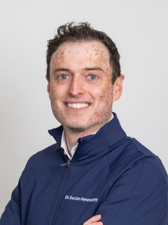 Smiling man with short curly hair and freckles wearing a navy blue jacket with embroidered text reading Dr. Declan Hennessey.