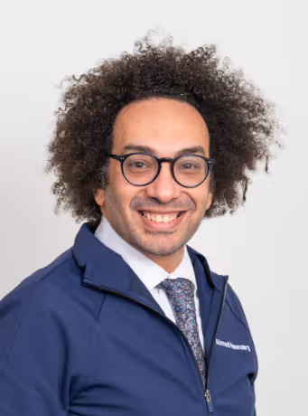 Smiling man with curly hair, glasses, wearing a navy jacket and patterned tie against a plain background.