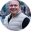 Smiling man wearing a light gray puffer vest and blue shirt outdoors in a city setting.
