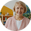 Smiling elderly woman with short blonde hair wearing a pink sweater and beige apron.