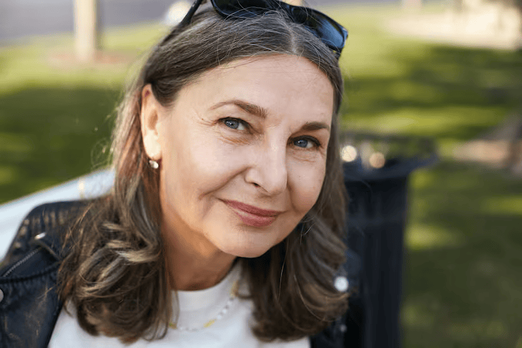 Close-up of a smiling middle-aged woman with gray hair outdoors, wearing sunglasses on her head.