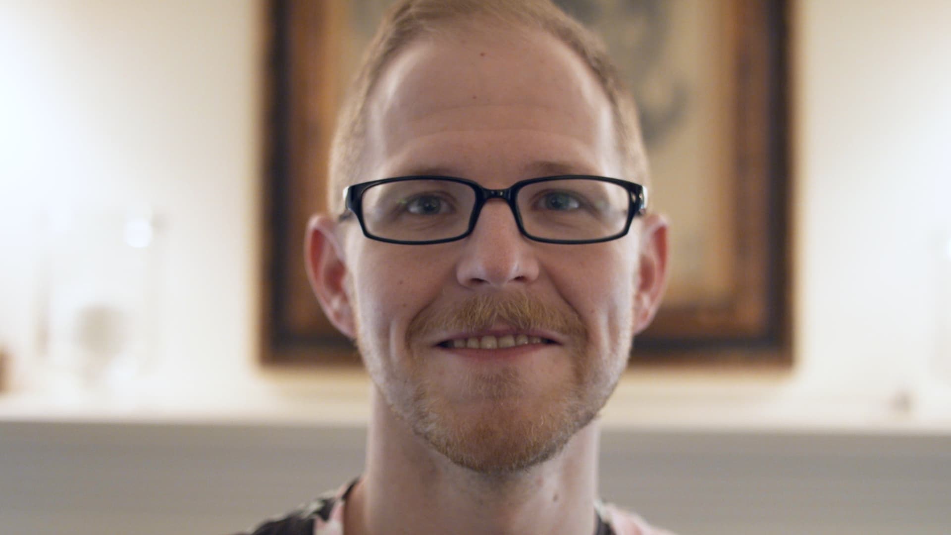 Smiling man with light beard and black glasses in a softly lit room with blurred background.