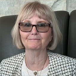 Portrait of an older woman with short blonde hair, glasses, and a black and white checkered jacket sitting on a dark chair.