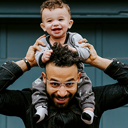 Smiling toddler sitting on an adult man's shoulders, both looking happy in front of a dark blue wall.