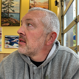 Middle-aged man with gray hair and beard wearing a gray hoodie, looking to the left indoors with framed landscape paintings on the wall behind him.