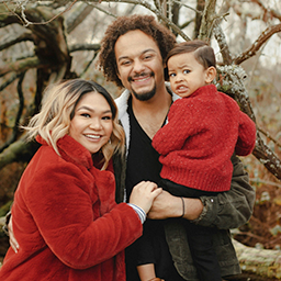 Happy family outdoors with a woman, man, and child wearing red sweaters, smiling near a tree.