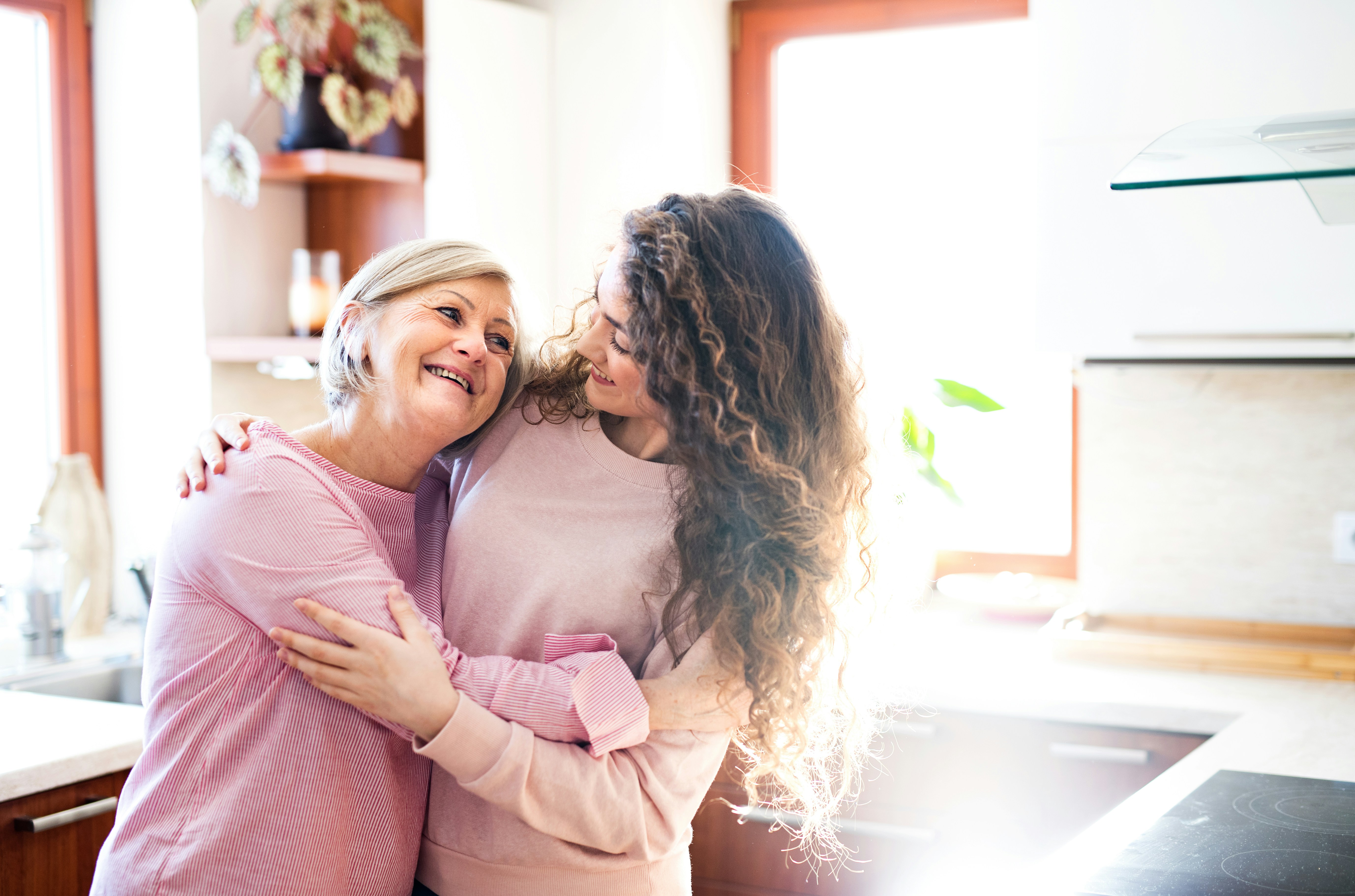 A mother and her grown daughter hugging in a sunny kitchen