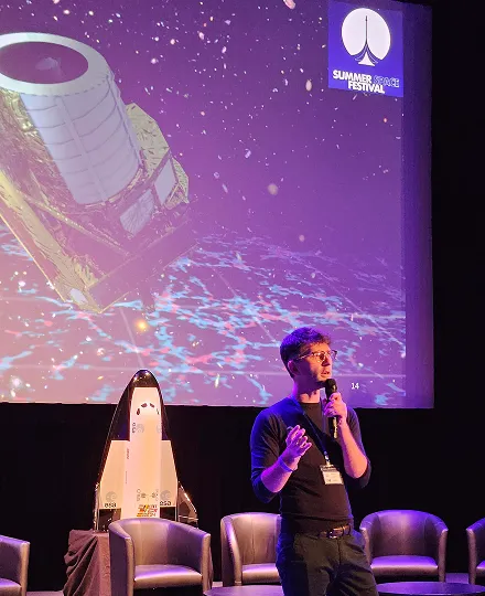 Speaker presenting at Summer Space Festival with a large screen showing a space telescope and a small space shuttle model on stage.