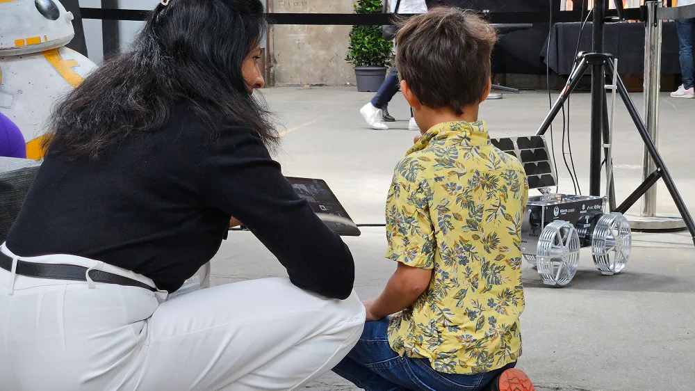 Woman and boy kneeling indoors observing a small robotic vehicle with large wheels at the Summer Space Festival.