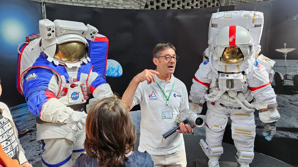 Man explaining features of two NASA astronaut suits to a group of people in an indoor exhibit at the Summer Space Festival.