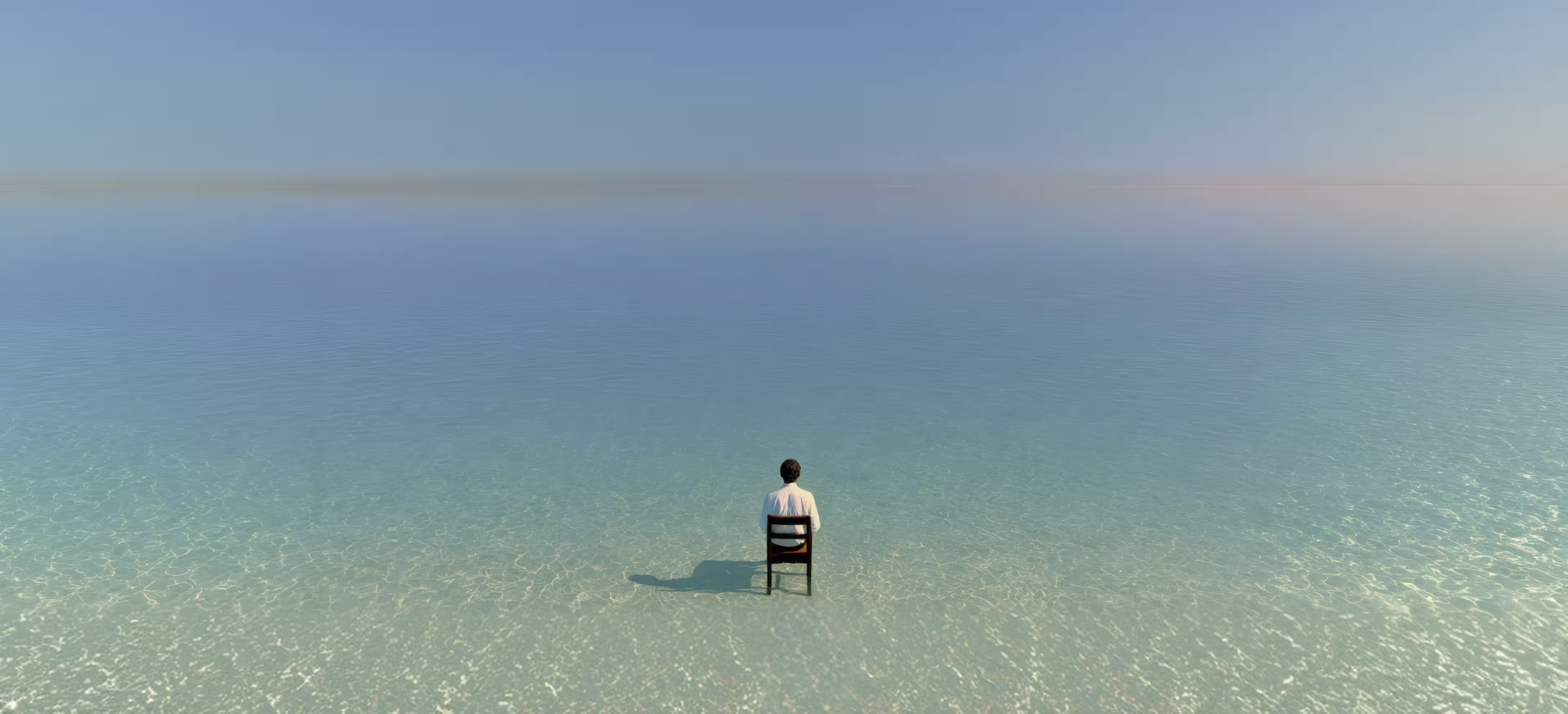 Person sitting on a single wooden chair in shallow clear water, facing away toward a calm, expansive ocean horizon under a clear sky.