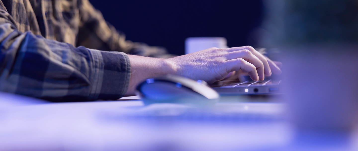 Person typing on a laptop keyboard wearing a plaid shirt with a wireless mouse nearby on the desk.