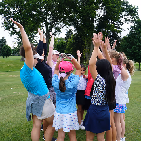 A group of girls and women on a golf course raising their hands in a cheer circle.