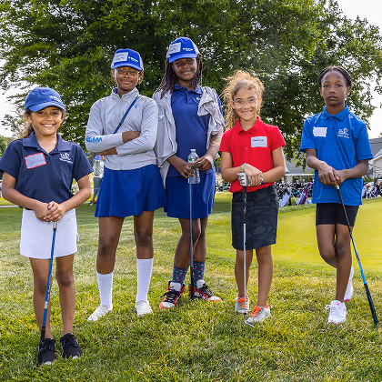 Five girls in golf attire standing on grass holding golf clubs, smiling at the camera outdoors.