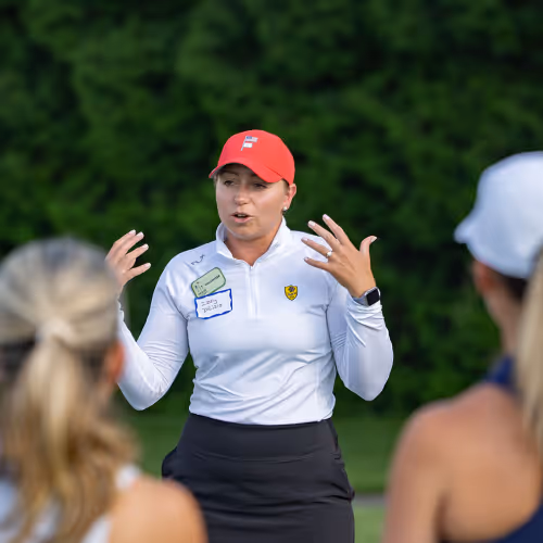 Woman in red cap and white shirt speaking to two people outdoors with green foliage background.