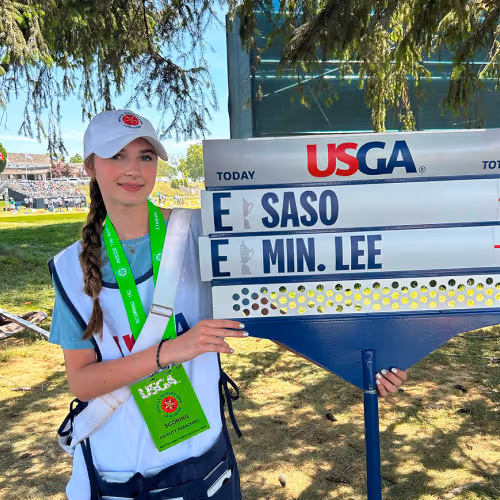 Young female USGA official holding a golf leaderboard showing SASO and MIN. LEE tied at even par.