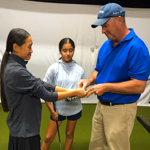 Man in blue shirt and cap teaching two young girls holding golf clubs indoors.