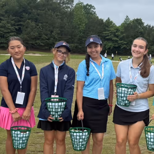Four girls standing on a golf course holding green baskets filled with golf balls, smiling for the camera.