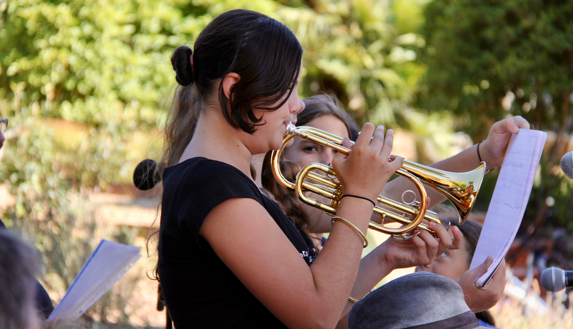 Une jeune fille en train de jouer de la trompette, avec une autre personne tenant une partition à côté d'elle, en extérieur.