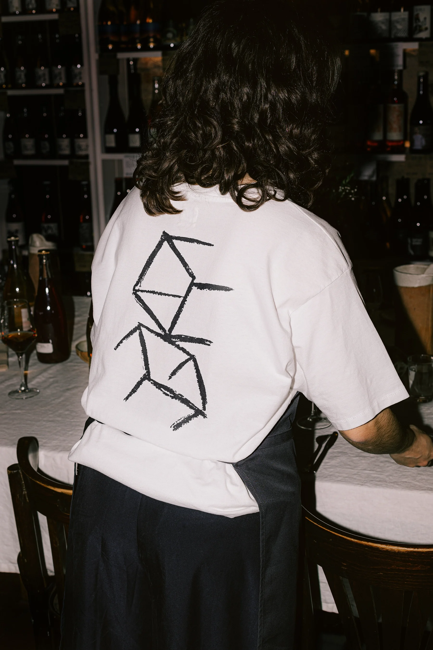 Person with curly hair wearing a white t-shirt with a black geometric design on the back and a dark apron, standing by a table with bottles and glasses.