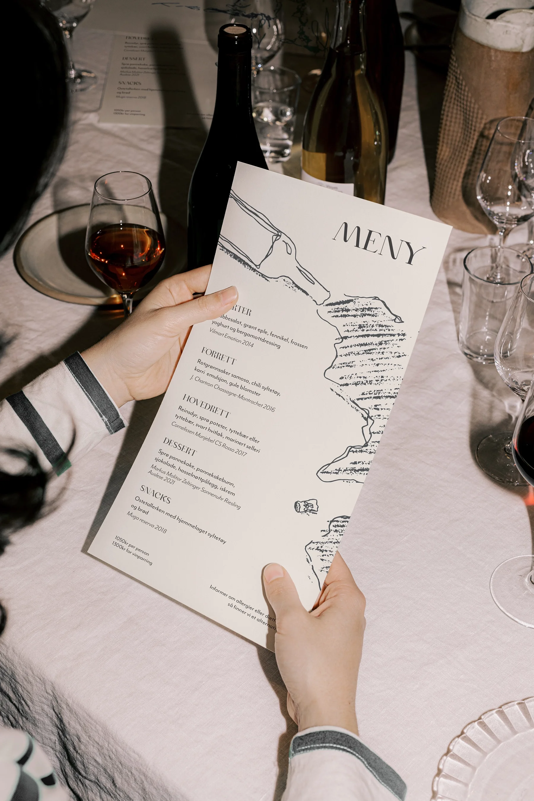 Person holding a menu titled 'MENY' at a table set with wine glasses, plates, and bottles.