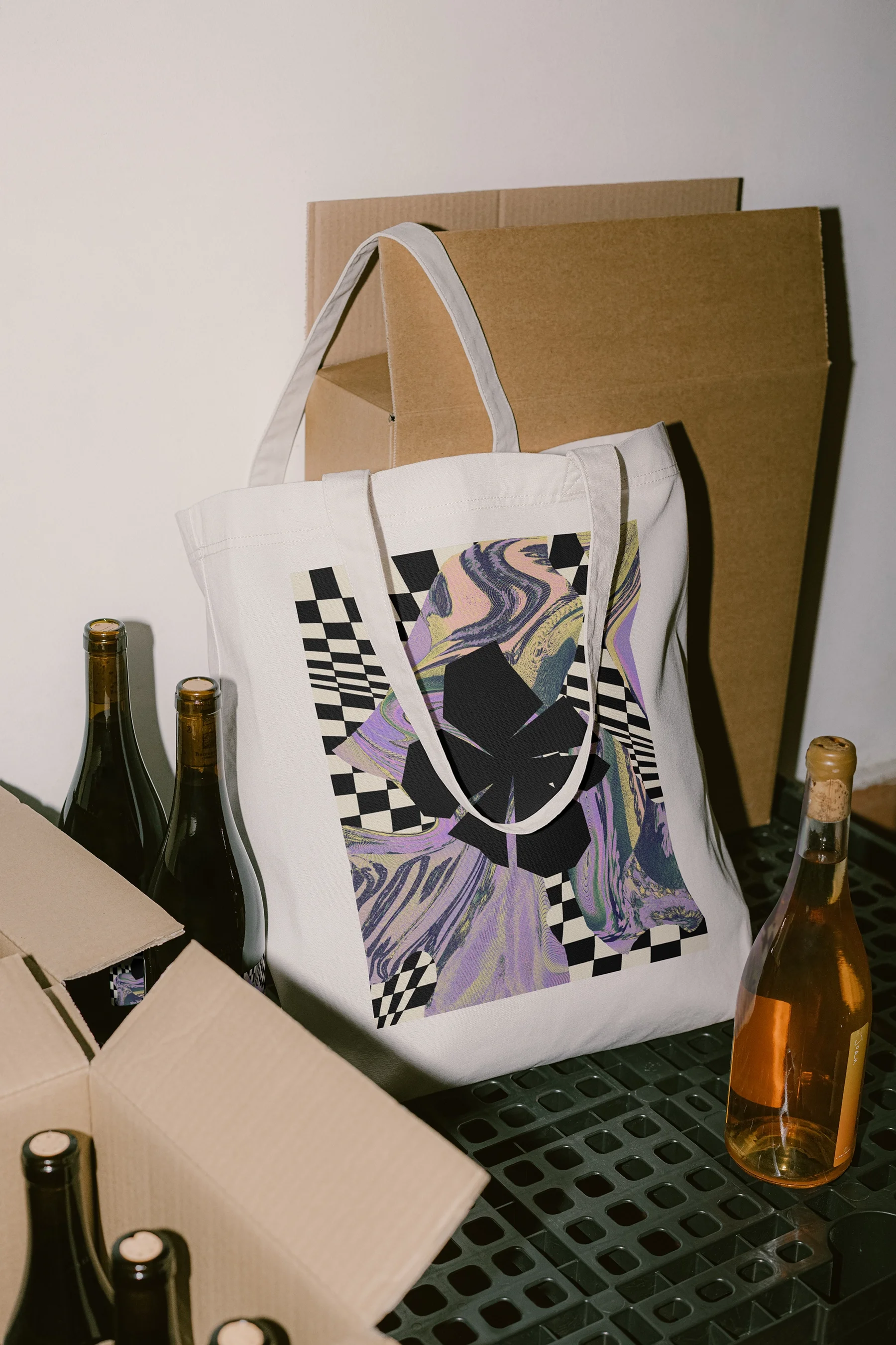 White tote bag with an abstract checkered and floral design surrounded by wine bottles and cardboard boxes.