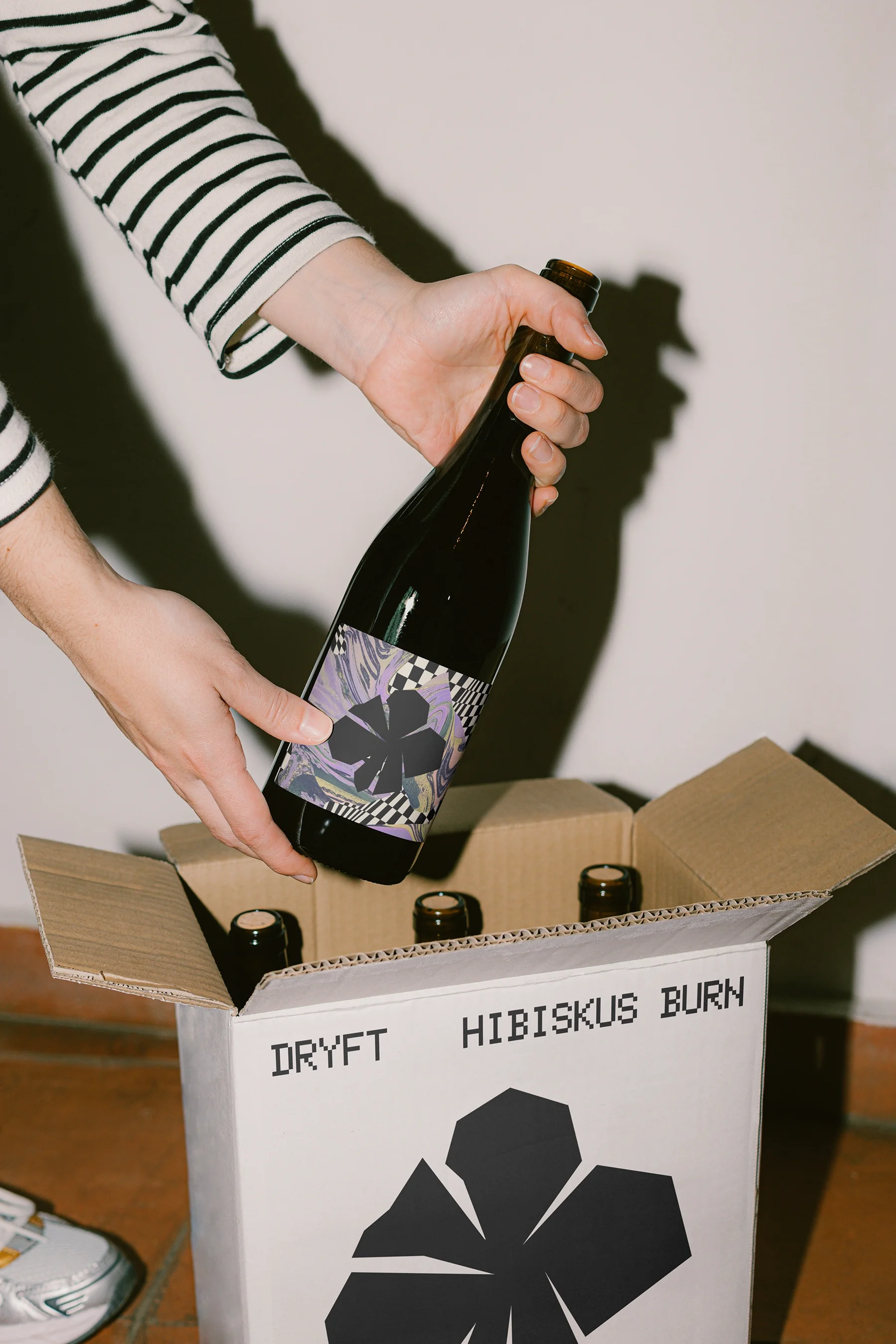 Person wearing a striped long-sleeve shirt holding a bottle with a purple label over a cardboard box labeled DRYFT HIBISKUS BURN containing more bottles.