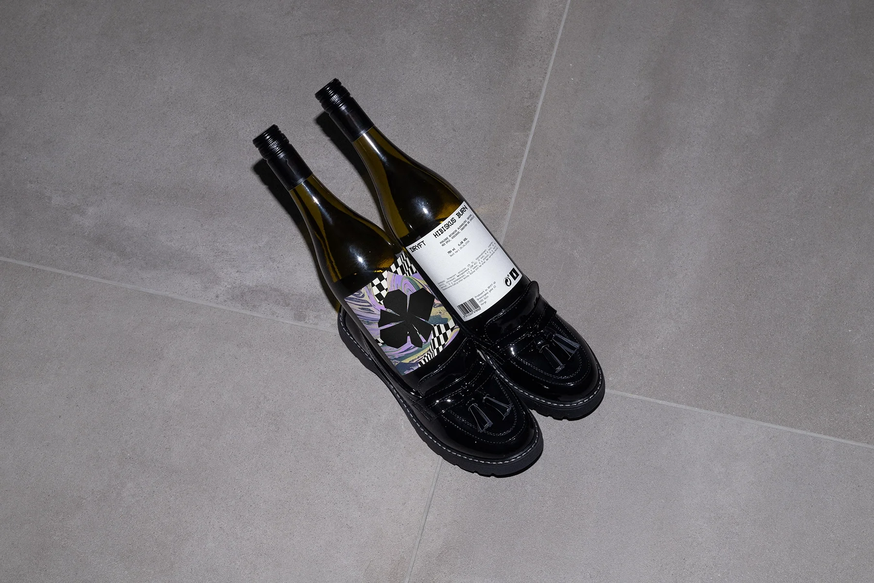 Two wine bottles placed inside shiny black tassel loafers on a light gray tiled floor.