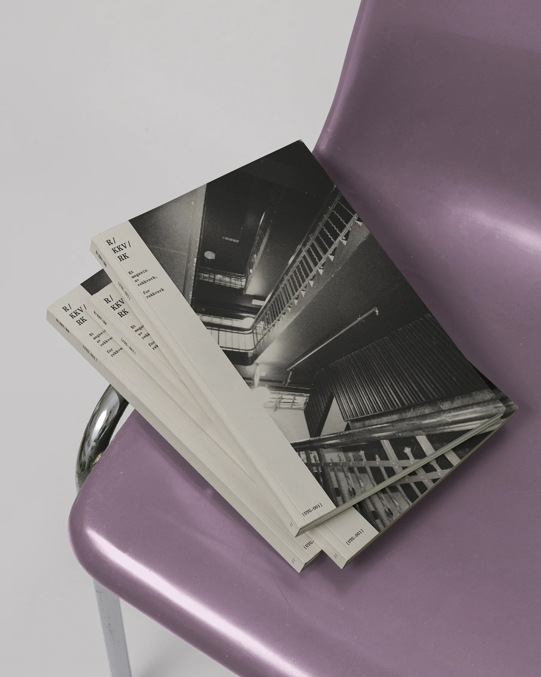 Four copies of a magazine titled 'R/KKV/RK' featuring a black-and-white photo of an industrial stairwell, placed on a purple plastic chair with a metal frame.