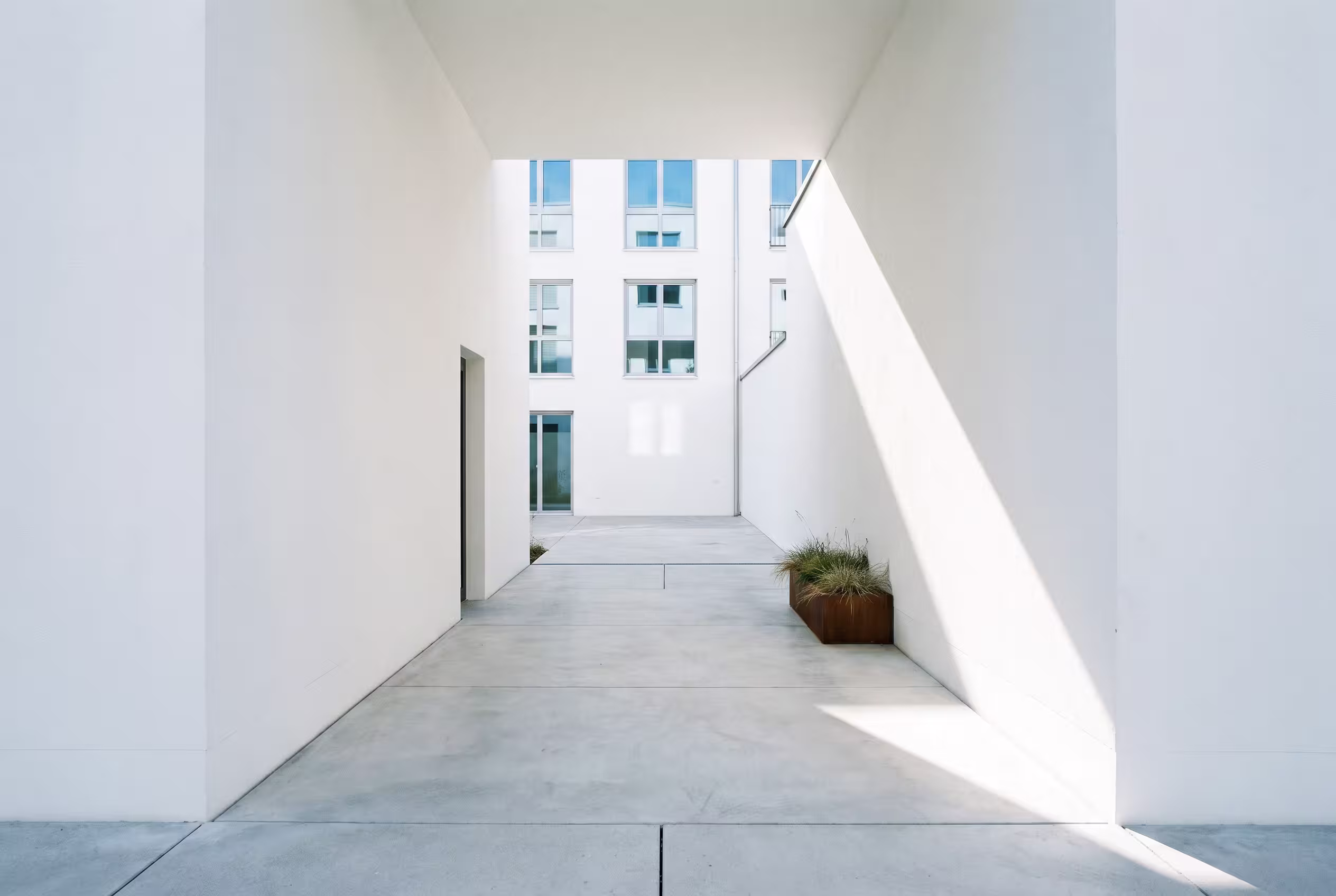 Minimalistischer weißer Gebäudekorridor mit rechteckigen Pflanzkübeln, die Licht- und Schattenmuster werfen.